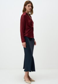Blusa di satin bordeaux con frontale attorcigliato e colletto, abbinata a una gonna satinata blu navy a campana e décolleté beige a punta.