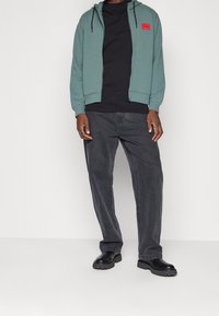 Zelená mikina na zip s černými doplňky, s červenou nášivkou loga, v kombinaci s černým tričkem a tmavě šedými džíny. Černé boty doplňují vzhled.