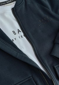 Jūras spēku zip-up kapuce ar gludu tekstūru un baltu T-kreklu zem tās, uz abiem apģērbiem ir melns teksts "BAKER BY TED BAKER".