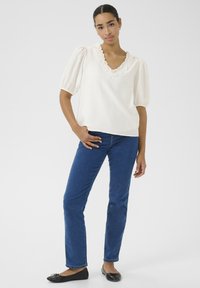 Blouse blanche avec un col en V volanté et des manches bouffantes. Associée à un jean droit bleu et des ballerines noires. Texture lisse et coupe décontractée.