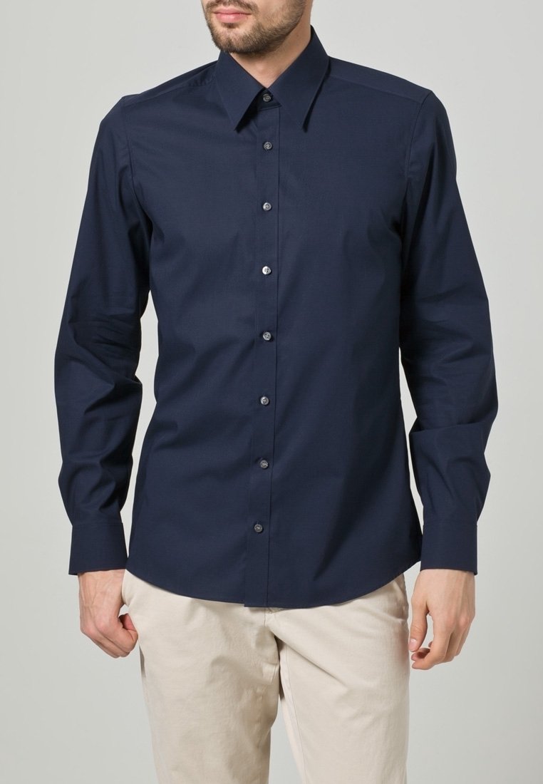 Homme portant une chemise bleu marine foncé à manches longues avec col et un pantalon beige clair sur un fond uni.