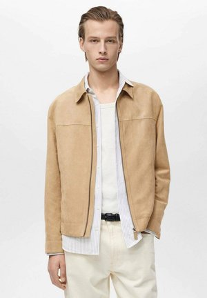 Giacca in suede beige chiaro con colletto, chiusura con zip completa e due tasche sul petto, indossata sopra una camicia bianca a righe blu.