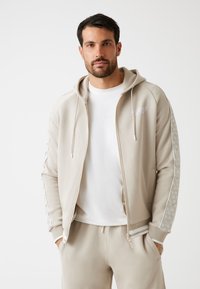 Beige zip-up hoodie met een trekkoordcapuchon, witte stiksels en een geautomatiseerd patroon aan de zijkanten. Gepaarde met een wit T-shirt en bijpassende broek.