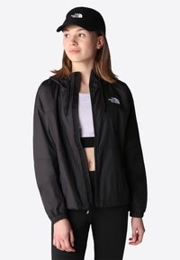 Svart lett zip-up jakke med elastiske mansjetter, med North Face-logoen på brystet og hette. Brukt over en sportstop.
