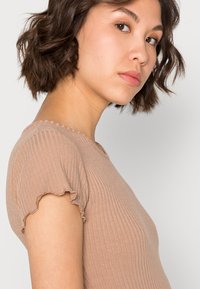 Top canela claro com nervuras, apresentando um design justo e mangas curtas com babados. Textura suave com um brilho subtil, decote redondo.