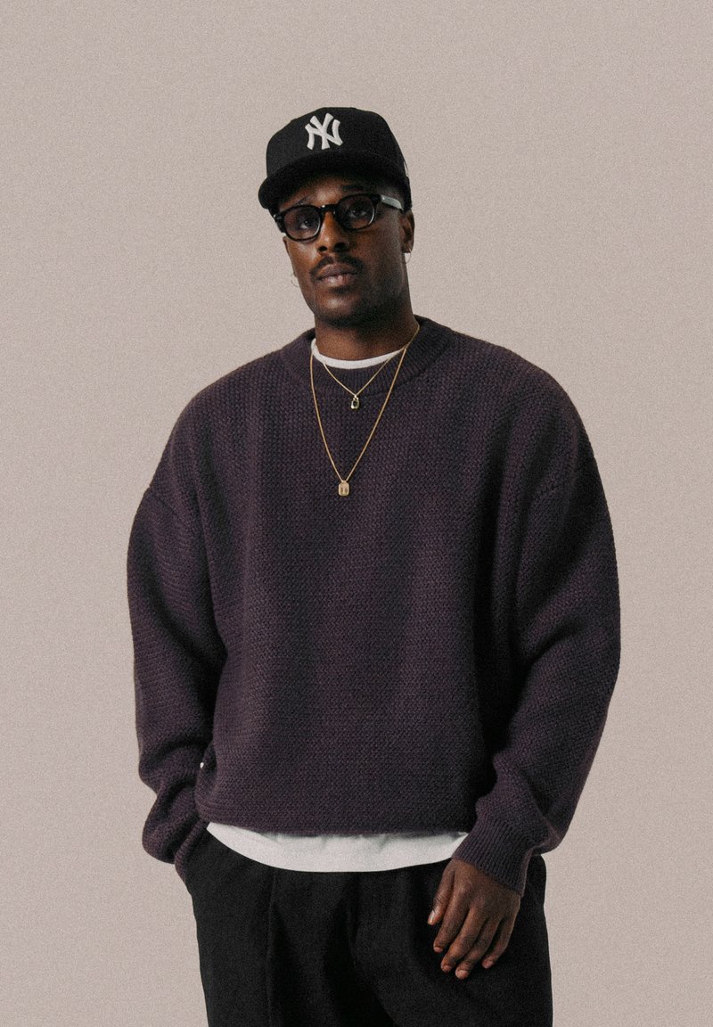 Lila Strickpullover mit strukturierter Oberfläche, runder Halsauschnitt, über einem weißen Shirt getragen. Kombiniert mit einer schwarzen Baseballkappe und Brille.