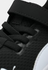 Chaussure de sport noire avec texture en maille, dotée de lacets plats, d'une bande auto-agrippante, et d'un accent brillant près de la zone des lacets.