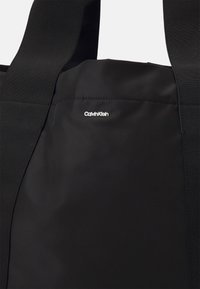 Svart tygkasse med breda remmar och en liten silverfärgad Calvin Klein-logotyp i mitten fram.