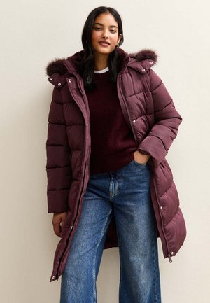 Burgundyröd puffjacka med huva i konstpäls, quiltad textur, framtida dragkedja och tryckknappsdetailler, bärs över en mörk tröja och vida jeans.