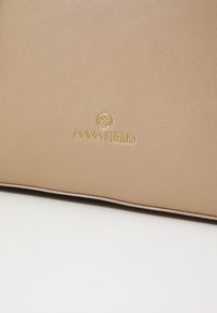 Beige læderhandtaske med prægede guld "ANNA FIELD" logo, glat tekstur, rene linjer og en nedtonet design. Ingen synlig hardware eller accenter.