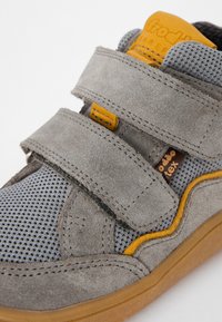Sandalie gri din suede cu panouri din plasă, având două curele Velcro galbene și o etichetă maro. Suprafață texturată și talpă din cauciuc.