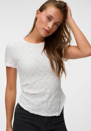 Jeune femme aux longs cheveux bruns portant un haut blanc texturé à manches courtes et un pantalon noir, touchant ses cheveux d'une main.