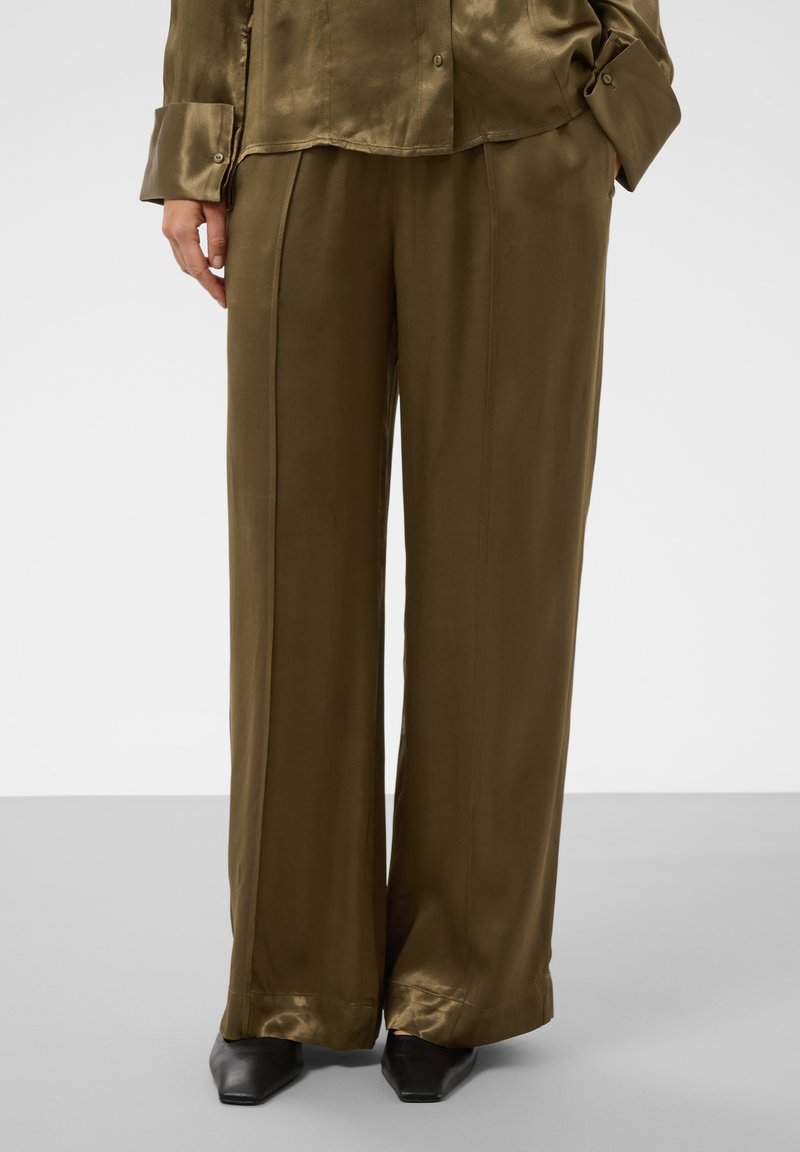 Personne portant un pantalon large en satin vert olive avec une chemise assortie et des chaussures noires à bout pointu, debout les mains dans les poches.