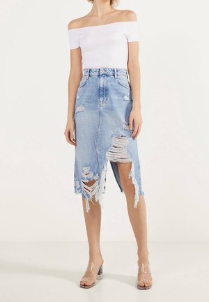 Vrouw draagt een witte off-shoulder top en een gescheurde lichtblauwe midi spijkerrok met hakken met doorzichtige bandjes.