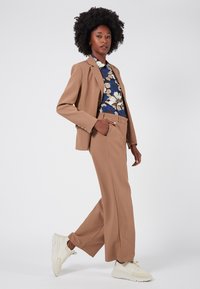 Beige op maat gemaakt pak met een getailleerde blazer en wijde broekspijpen, gecombineerd met een bloemenpatroon shirt. Witte sportschoenen maken de look compleet.