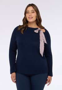 Maglione blu navy con orlo e polsini a costine, presenta una spalla ritagliata e un accento a cravatta bianca e rossa con motivo sul collo.