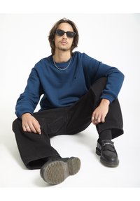 Blauwe sweatshirt, zwarte baggy broek, zwarte instappers en zonnebril. Zilveren ketting. Model zit met de handen op de knieën.