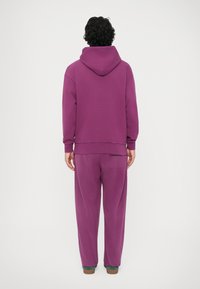 Hanorac mov cu glugă, având o croială lejeră. Asortat cu pantaloni de trening largi, de aceeași culoare. Textură moale, design simplu.