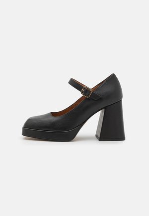 Chaussure à talon haut en cuir noir avec bout rond, bride Mary Jane et talon carré, présentant une surface texturée et un détail de boucle métallique.