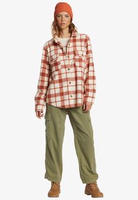 Checked flannel shirt in orange and cream, featuring button front, two chest pockets, and worn with olive green cargo pants and brown boots.