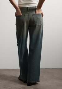 Persona vistiendo jeans de mezclilla oscura y holgados, con las manos colocadas de manera casual en los bolsillos traseros, de pie sobre un suelo gris contra una pared blanca.