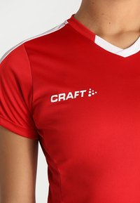 Rød sportsjersey lavet af glat stof, med hvid krave og skulderdetaljer, og med "CRAFT" logoet trykt i hvid på venstre side.