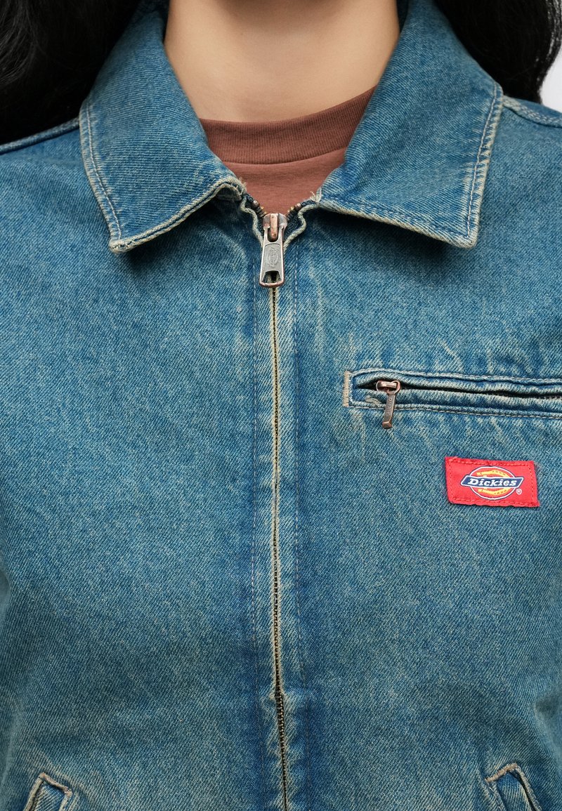 Spijkerjack dichtgeritst over een bruin shirt, met een borstzak met rits en een rood Dickies-merkpatch aan de rechterkant.