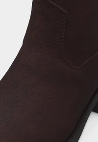 Κοντινό πλάνο σκούρου καφέ μποτάκι από σουέτ με ελαστικό πλαϊνό πάνελ και χαμηλό μαύρο τακούνι.