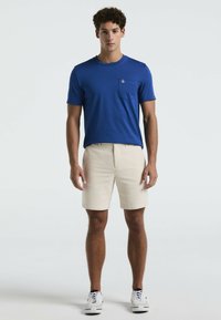 Chemise bleu à manches courtes avec poche, shorts beiges et baskets blanches. Tissu lisse, coupe décontractée, design simple, adapté pour un usage décontracté.