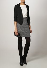 Blazer noir à manches trois-quarts, blouse crème, jupe grise texturée, collants noirs et bottines. Tient une pochette noire et argent.