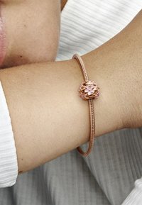Braccialetto rigido in oro rosa con un ciondolo a forma di fiore, caratterizzato da dettagli in smalto rosa. Texture metallica liscia, design flessibile.