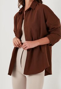 Donna che sbottona una camicia a maniche lunghe marrone scuro sopra un top beige e pantaloni beige su uno sfondo semplice.