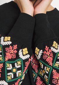 Sudadera negra con bordados florales coloridos en rojo, amarillo, verde y blanco en las mangas, con puños acanalados y patrones detallados.
