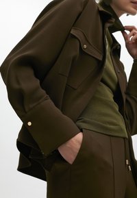 Giacca marrone con un design strutturato, dettagli in metallo dorato e tasche laterali, indossata sopra un top verde scuro con texture a coste.