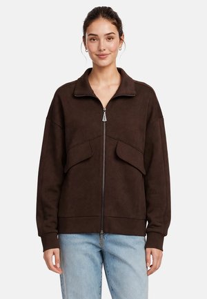 Donna che indossa una giacca marrone scuro con zip e tasche con patta e jeans azzurri, in piedi davanti a uno sfondo semplice.