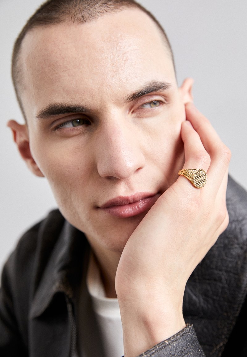 Zlati prstan z teksturirano površino, nošen na roki, ki počiva na obrazu, na nevtralni podlagi. Poudarek na tonih kože in podrobnostih prstana.