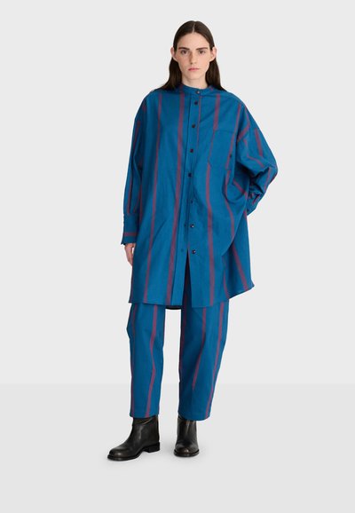 Femme portant une tenue bleue ample avec des rayures verticales bordeaux, une longue chemise boutonnée, un pantalon assorti et des bottines noires sur fond uni.