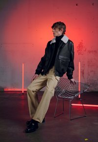 Black leather bomber jacket with white fur collar, paired with tan wide-leg pants. Model sits on a wire chair against a red backdrop.