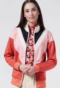 Färgblockerad zip-up jacka med paneler i korall, rosa och svart. Inuti finns en blommig mock neck-tröja och ribbade mudd vid ärmarna. Slät textur.