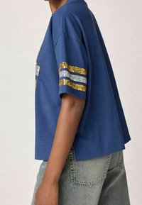 Camiseta de manga corta azul marino con rayas de lentejuelas doradas y plateadas en las mangas. Corte holgado, fabricada en una tela suave con una textura lisa.