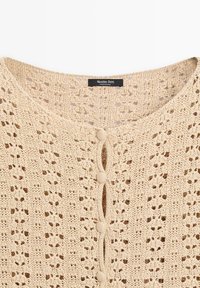 Cardigan beige en tricot crocheté avec col rond, boutons recouverts de tissu et étiquette Massimo Dutti à l'arrière du col.