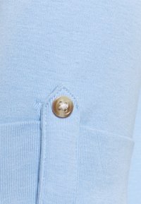 Light blue knitted fabric with a textured finish, featuring a gold button and an angled placket at the side.