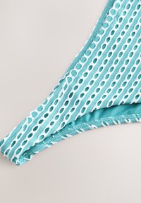 Turkio spalvos maudymosi kostiumėlis su išpjovomis ir baltais akcentais. Turi lygią tekstūrą ir siūtas kraštines, užtikrinančias saugų prigludimą.