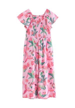 Smilende ung pige med krøllet hår iført en pink kjole med blomster- og fuglemønster og brune flettede sko, stående mod en ensfarvet hvid baggrund.