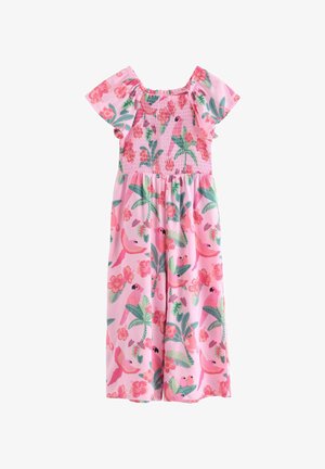 Smilende ung pige med krøllet hår iført en pink kjole med blomster- og fuglemønster og brune flettede sko, stående mod en ensfarvet hvid baggrund.