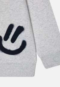 Sudadera gris con un gran gráfico de cara sonriente azul marino en la manga, hecha de algodón suave con un acabado texturado similar al forro polar.