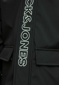 Giacca nera con logo "CK&JONES" bianco sul lato, con una texture liscia e opaca e due tasche frontali a soffietto.