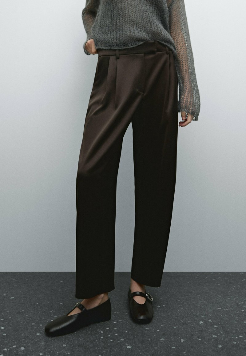 Brown, high-waisted, pleated trousers with a fluid design. Paired with black slip-on shoes featuring a metallic accent. Textured gray sweater.