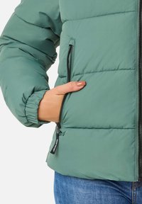 Zielona kurtka ocieplana z wysokim kołnierzem, kieszeniami na zamek, elastycznymi mankietami i teksturowaną powierzchnią. Widoczne linie szwów podkreślają projekt.