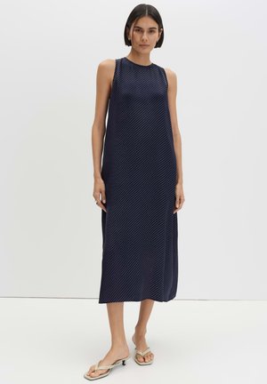 Vrouw draagt een mouwloze marineblauwe midi-jurk met diagonale strepen en beige open-teen sandaaltjes met hak, staand tegen een effen achtergrond.